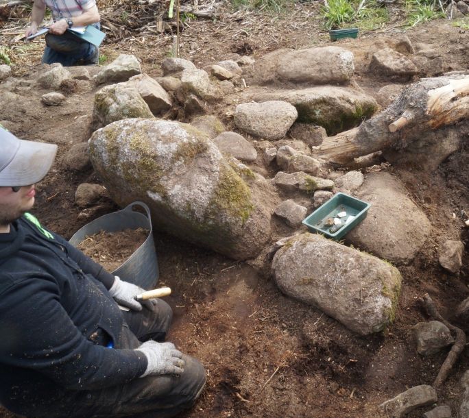Lewis finding a spindle whirl behind a large stone in front of him.